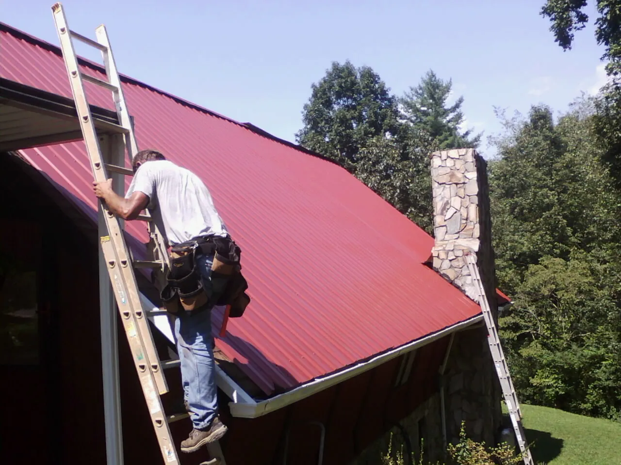 Roofer on a ladder beside a red metal roof and stone chimney — Soffit & Fascia Repair by Roofing Brothers in Patuxent River